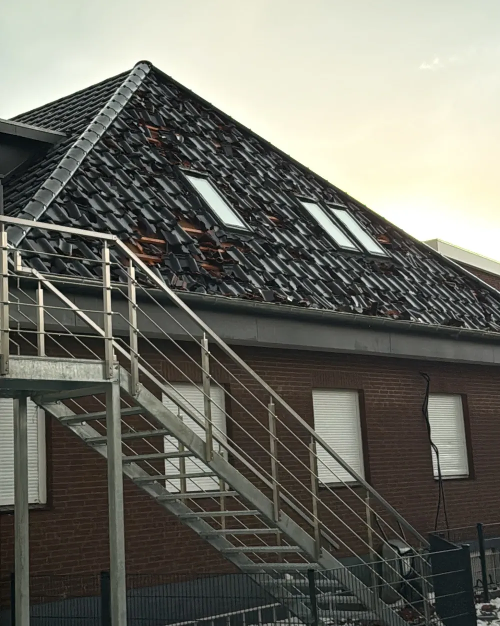 Klinkerhaus mit Außentreppe, auf dessen Dach viele Ziegel zerschlagen sind; darunter ist die Lattung zu sehen